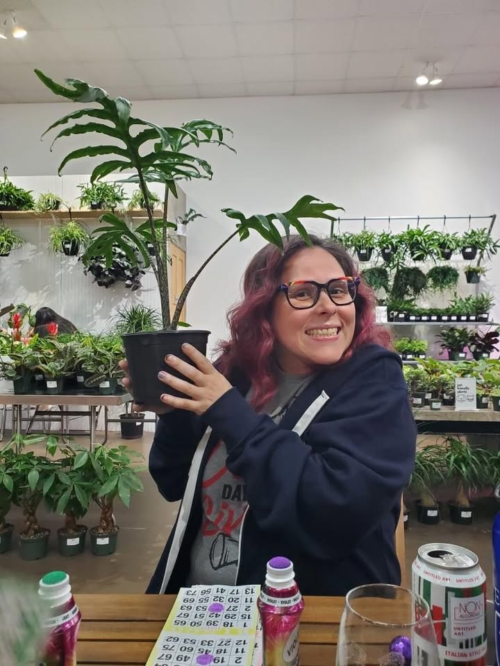Your plant lady, grinning and holding up a tall potted plant at a plant shop